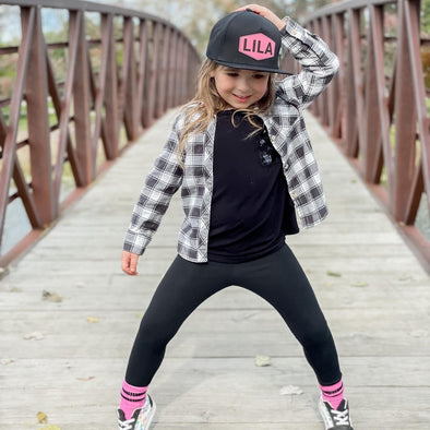 Shop online for snapback hats from LB Threads for baby, toddler, kid and adult. Toddler model wearing the LB Threads Malakai Custom Name hat, a black flat bill classic snapback hat with a pink patch with her name, LILA, on it.
