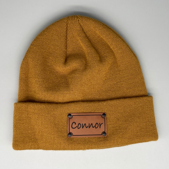 Shop online for beanies and snapback hats from LB Threads for babies, toddlers, kids and adults. Photo shows our Tan Custom Name Beanie, a warm, soft, tan acrylic beanie with a leather engraved custom name patch attached with rivets.