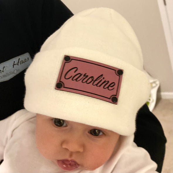 Shop online for beanies and snapback hats from LB Threads for babies, toddlers, kids and adults. Infant model wearing our Cream Custom Name Beanie, a warm, soft, cream-colored acrylic beanie with a leather engraved custom name patch attached with rivets.